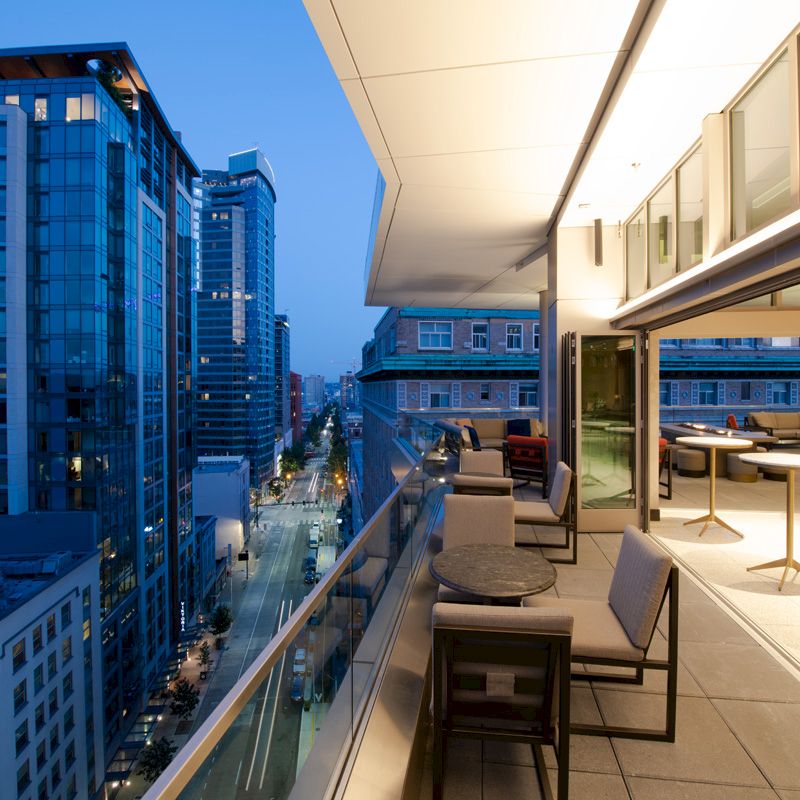 An outdoor balcony at dusk overlooks a city street and high-rise buildings, with tables and chairs lining the balcony.