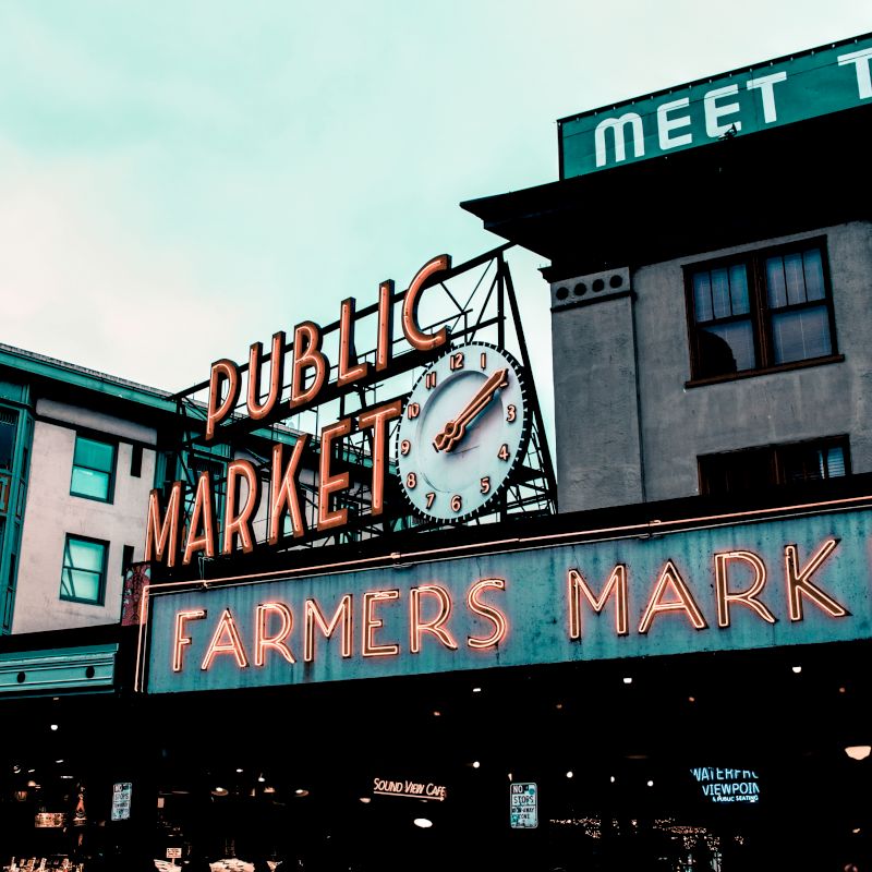 This image shows the iconic sign of a public market with a clock, reading "FARMERS MARKET" in neon letters, likely representing a popular city location.