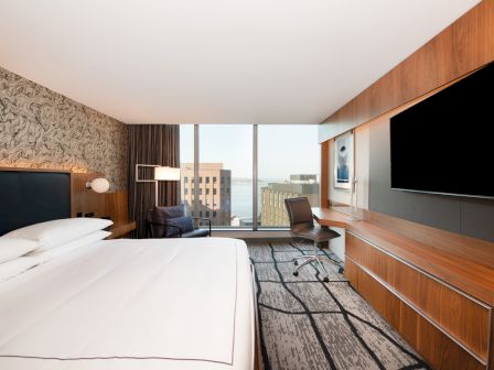 A modern hotel room with a large bed, desk, chair, flat-screen TV, and a window providing a view of buildings and a body of water.