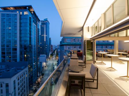 A modern cityscape at dusk, with tall buildings and a rooftop terrace featuring tables, chairs, and lounge areas, overlooking a lit street below.