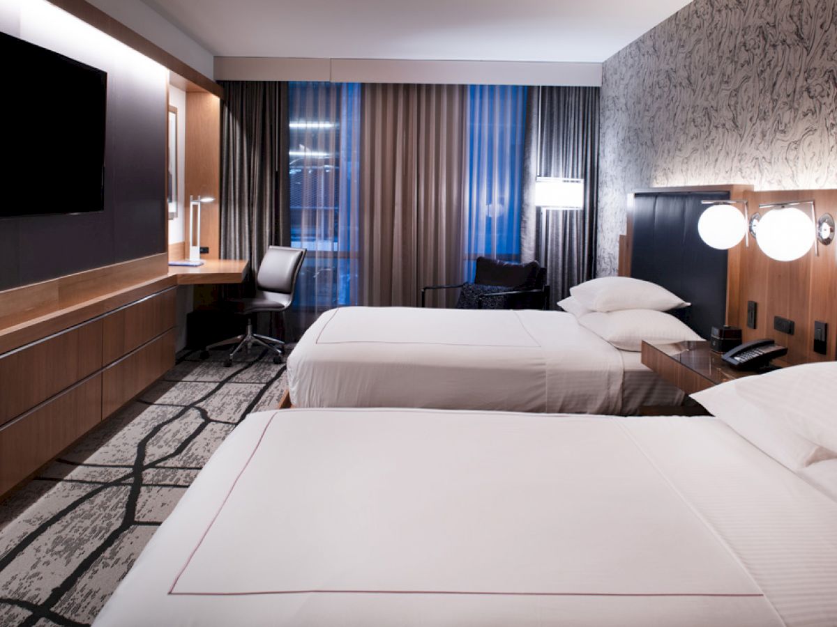 A modern hotel room with two beds, a wall-mounted TV, a desk by the window with a chair, and stylish lighting and decor, appearing clean and inviting.