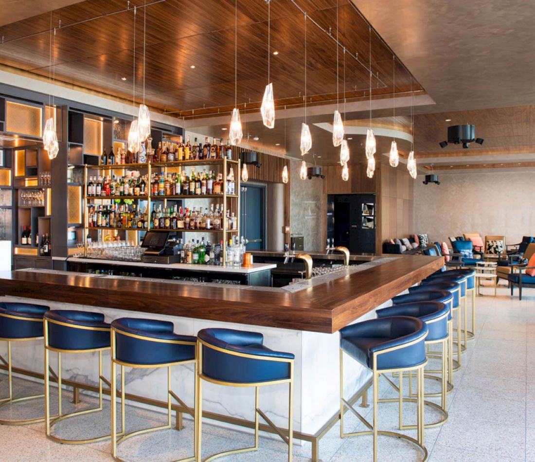 A wide view of a bar with a long counter, blue stools, and shelves of liquor behind the counter.