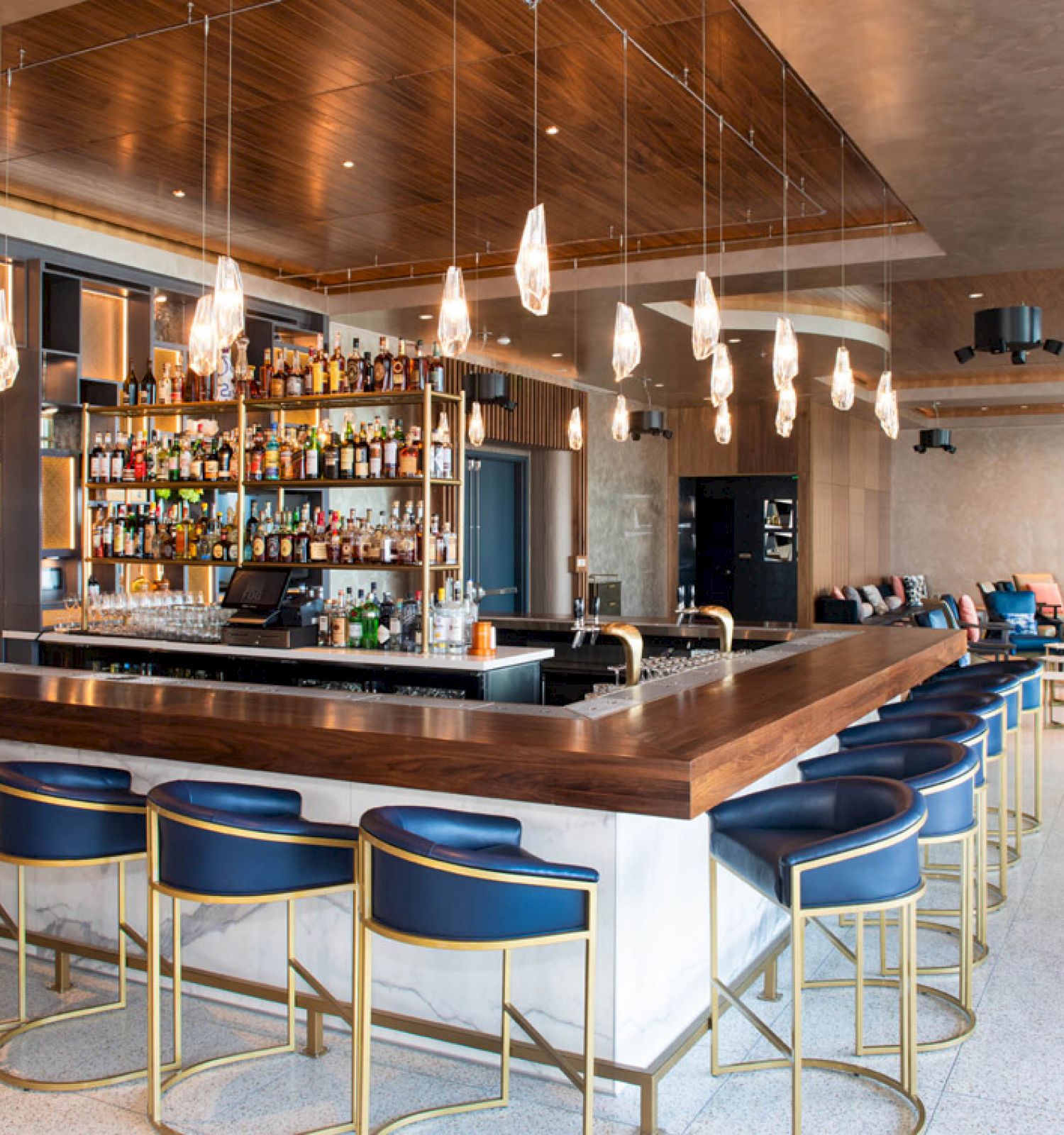 A modern bar with blue stools, wooden countertops, hanging lights, and a wide selection of bottles. The seating area is visible in the background.