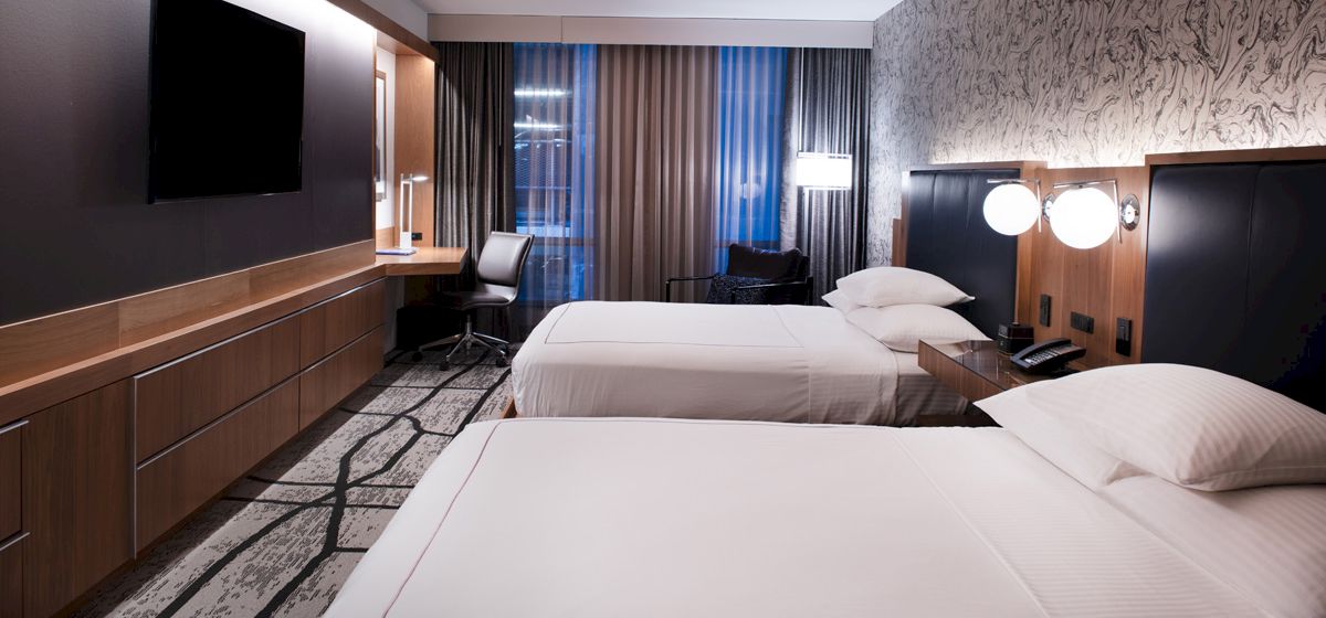 The image shows a modern hotel room with two neatly made beds, a wall-mounted TV, a desk and chair, and contemporary decor.