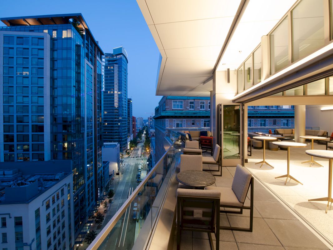 A modern cityscape at twilight, with a view of high-rise buildings and a stylish, open-air balcony featuring tables and chairs.