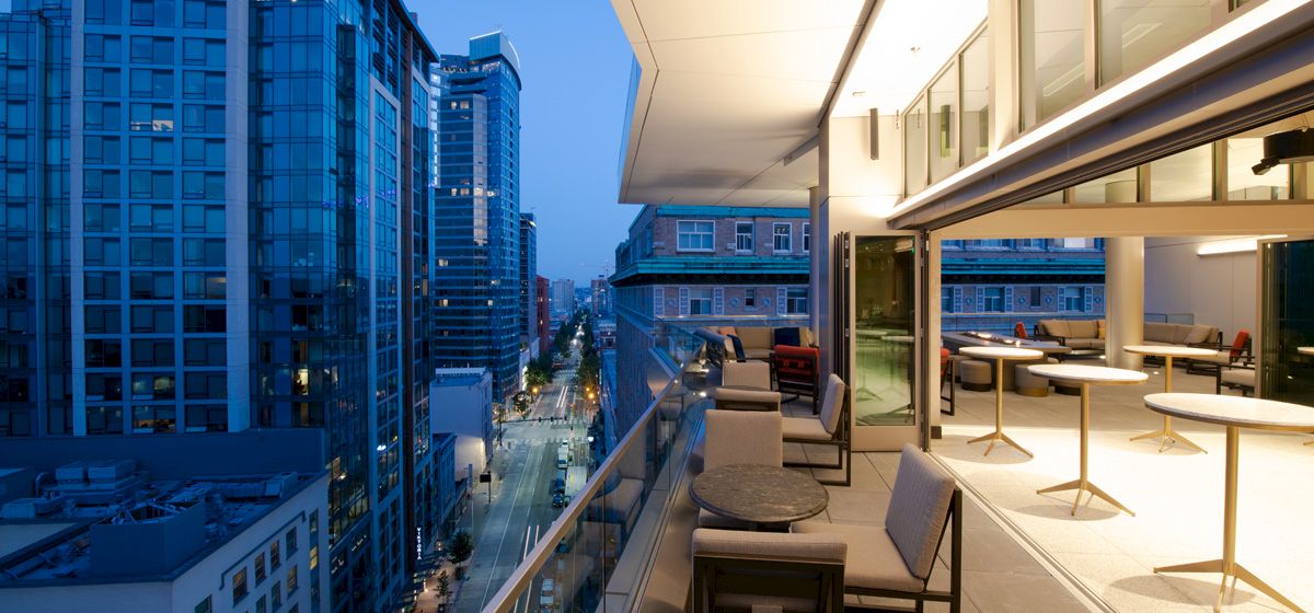 A modern cityscape at twilight, with a view of high-rise buildings and a stylish, open-air balcony featuring tables and chairs.