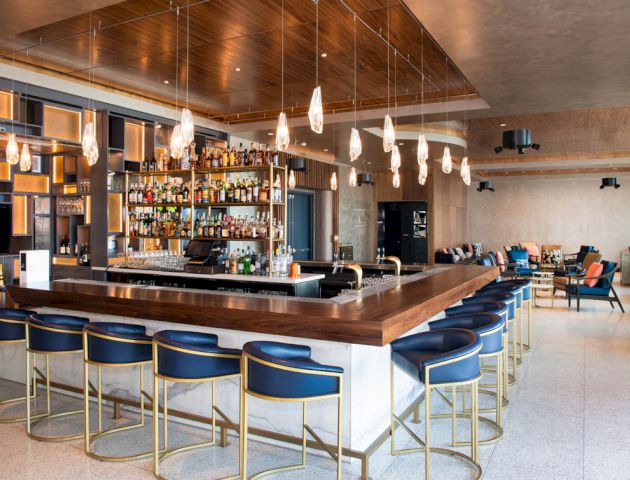 This image shows a modern bar with a wooden counter, blue bar stools, hanging lights, and shelves stocked with various bottles.