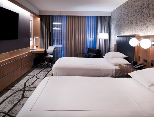 A modern hotel room with two neatly made beds, a TV, a desk, and large windows with curtains.