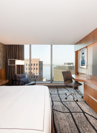 A modern hotel room with a large bed, wall-mounted TV, desk, chair, and a window with a city and water view.