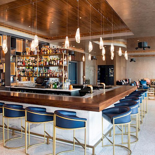 The image shows a modern bar with a wooden counter, blue barstools, multiple hanging lights, and a well-stocked back bar. The seating area is in the background.