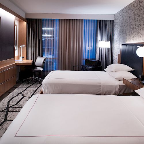 The image shows a modern hotel room with two beds, a flat-screen TV, a desk, and large windows with patterned curtains, offering a stylish and cozy ambiance.
