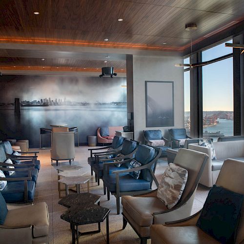 A stylish lounge with modern seating, large windows with a city and water view, and wall art depicting a skyline.
