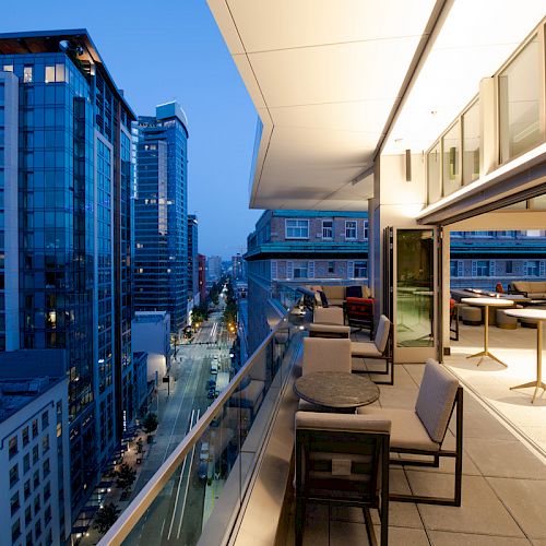 The image shows a rooftop balcony overlooking a cityscape at dusk, with modern buildings, tables, chairs, and an illuminated interior space.