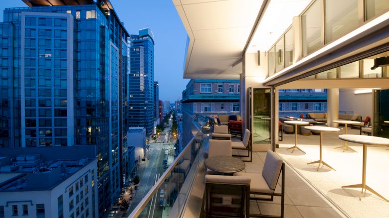 An urban balcony scene at dusk with modern buildings, furniture, and a glass railing overlooking a city street surrounded by tall structures.