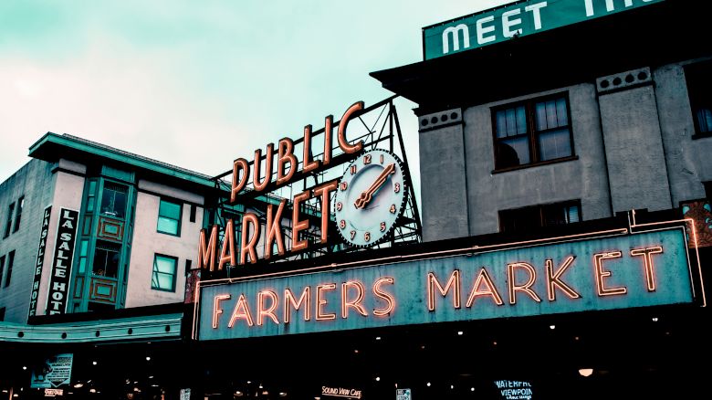 This image showcases the iconic neon sign of a public farmers market in an urban setting, with buildings in the background.