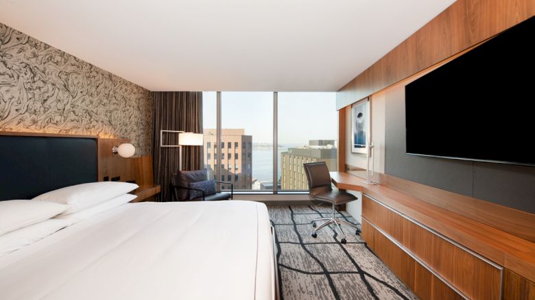 The image shows a modern hotel room with a large window, king-size bed, desk, flat-screen TV, and an urban view.