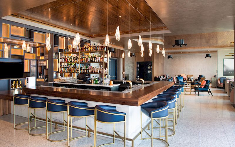 A modern bar with a central counter, blue stools, hanging lights, and a fully stocked shelf of liquor bottles in a spacious, stylish setting.
