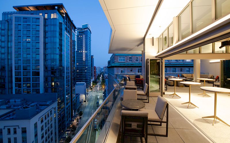 A modern balcony view of a cityscape at dusk, with tall buildings, a street below, and outdoor seating arrangements on the balcony.