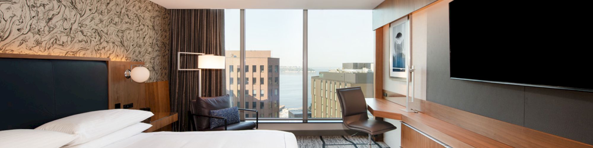 A modern hotel room features a large bed, a wall-mounted TV, a desk, and a window with a city view.