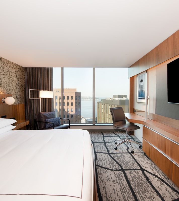 A hotel room with a bed, desk, chair, and TV, overlooking the city.