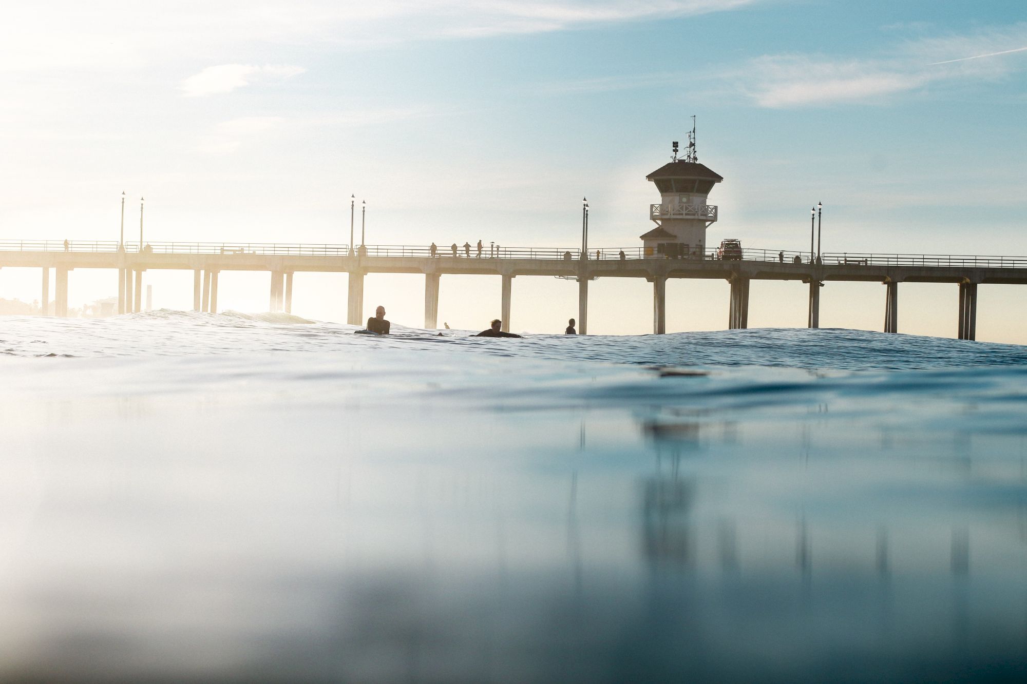 The image shows a view of a pier with people walking on it, seen from the water, with a few swimmers or surfers in the foreground.
